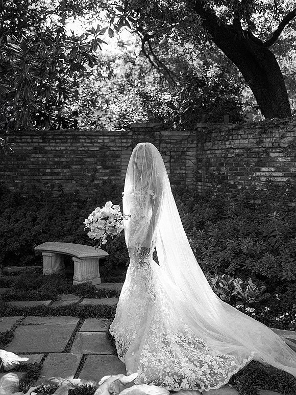 Bridal portrait of a bride in a lace wedding dress with a long veil over her face, holding a bouquet on a garden stone path by a brick wall