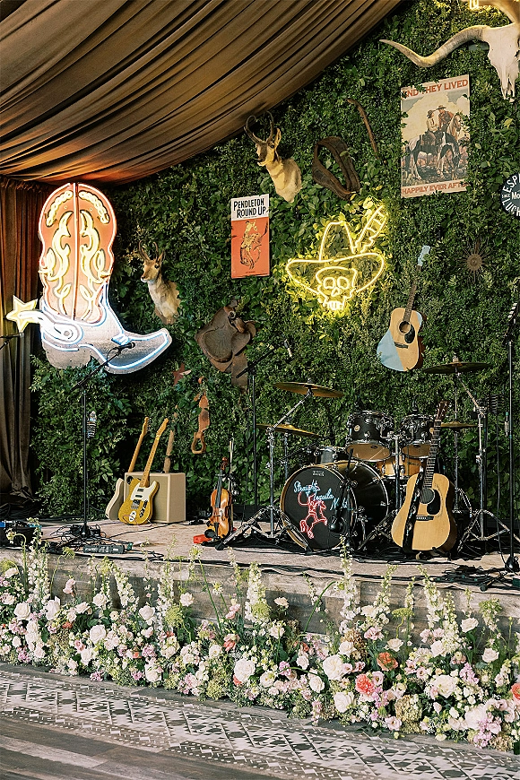 Wedding band stage with greenery wall backdrop, neon boot sign, drum kit, guitars, and flower-lined edge on an indoor dance floor