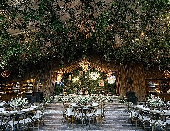 Reception decor with greenery ceiling wedding accents, string lights and chandeliers over round tables with candles and floral centerpieces in tent