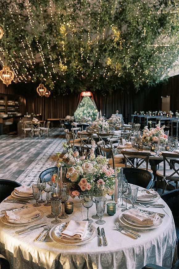 Reception tablescape with blush rose centerpiece and taper candles on a white linen round table under string lights and hanging lanterns