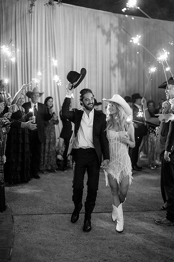 Wedding send off with a sparkler send off tunnel as the newlyweds in cowboy hats walk hand in hand past guests under draped fabric at night