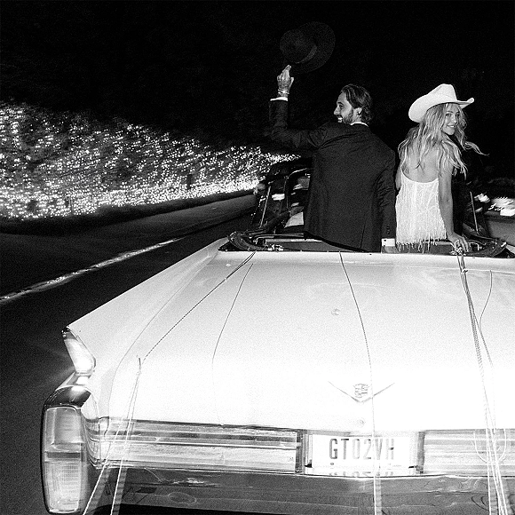 Wedding send off with newlyweds in convertible under string lights, groom waving cowboy hat beside bride in strapless dress on road at night