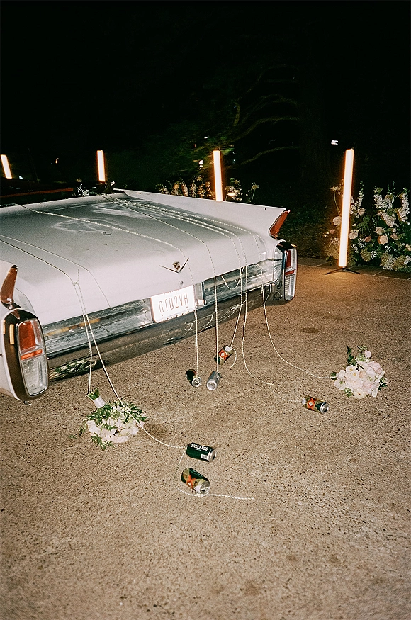 Wedding getaway car, a white vintage car with trailing ribbons and beer cans, taillights glowing in a nighttime garden driveway
