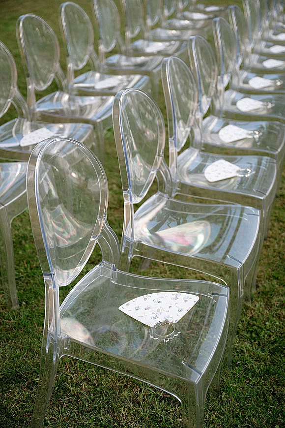 Ceremony seating with clear acrylic wedding chairs arranged in rows on a grass lawn, each seat topped with a paper fan for guests