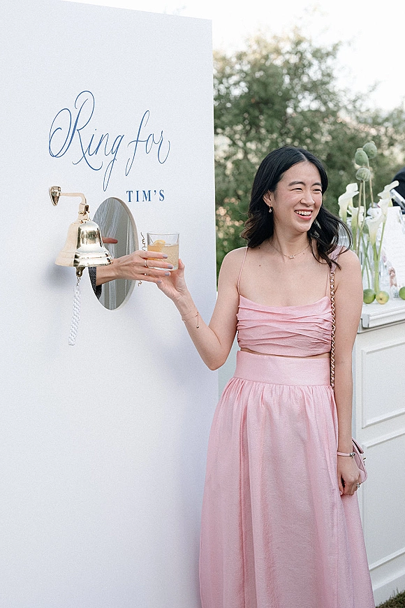 Wedding cocktail moment as a guest in a pink satin dress holds a drink by a wedding drink wall with a service bell and calligraphy sign outdoors