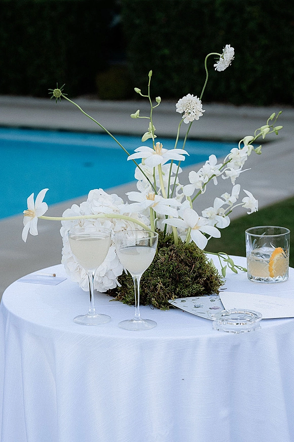Reception tablescape with wedding table centerpiece of white flowers and moss on white linen, wine glasses and citrus cocktail by the pool