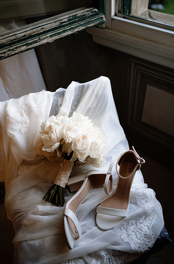 Wedding details flat lay with bridal shoes and bouquet on a lace wedding dress draped over a distressed wooden chair in window light