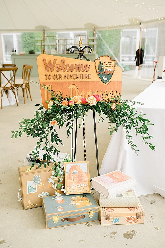 Wedding welcome sign on a metal easel with greenery garland and pink-peach roses, framed by travel suitcases and string lights in a white tent