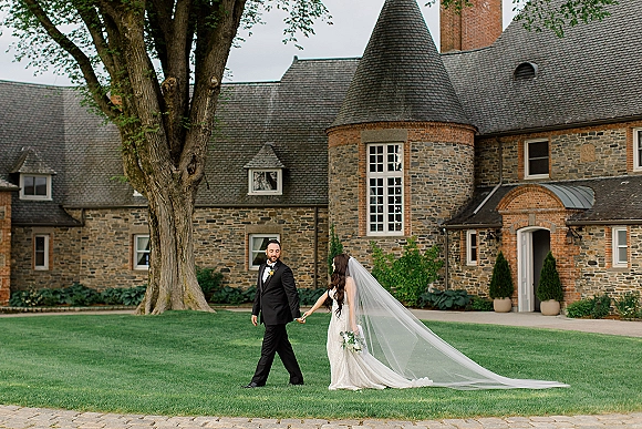 Couple portrait of bride and groom holding hands, walking on an estate lawn with bride’s veil flowing, stone manor behind