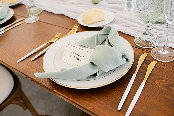 Wedding place setting with a sage green napkin knot, place card on plate, gold and white flatware, goblet and wine glass on wood table
