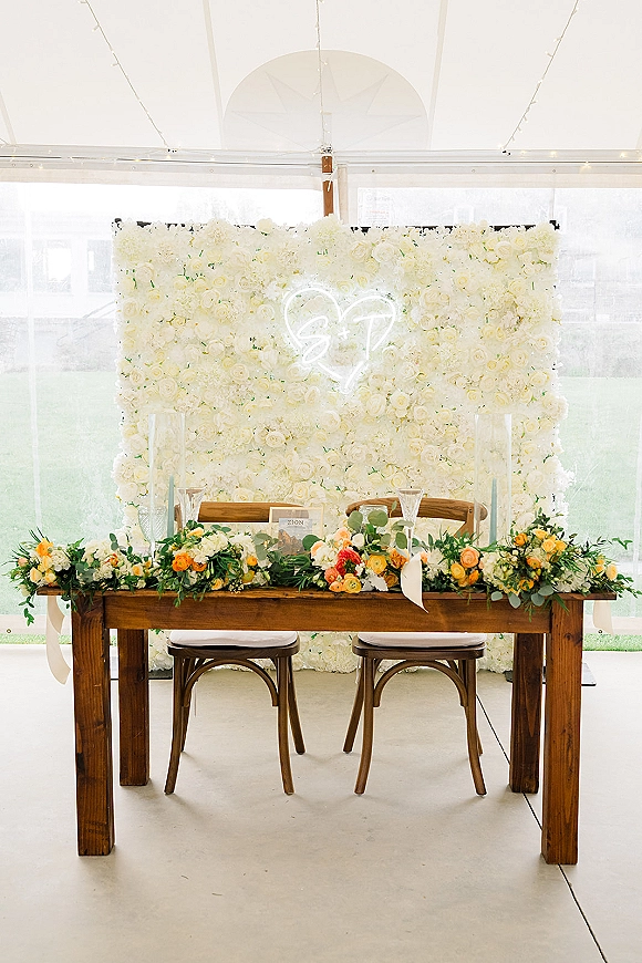 Reception sweetheart table with a white flower wall and neon heart sign, greenery garland, candles, and place settings in a clear tent with string lights