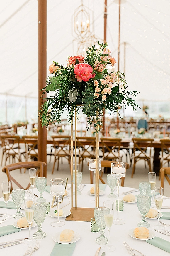 Reception tablescape with tall floral centerpiece on a gold stand, coral peonies, candles, mint napkins, in a clear tent with string lights