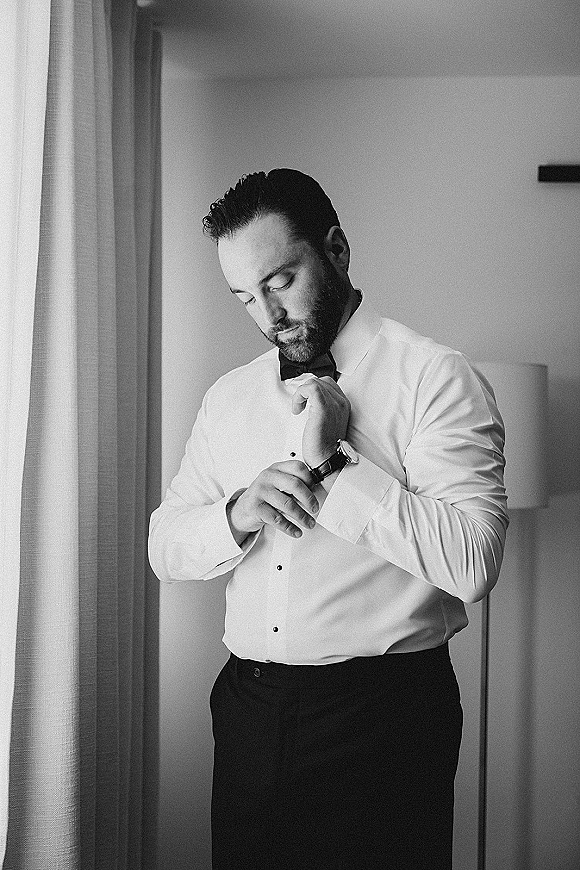 Groom getting ready adjusts his bow tie and checks his wristwatch by a window with curtains and a lamp, wearing a white dress shirt