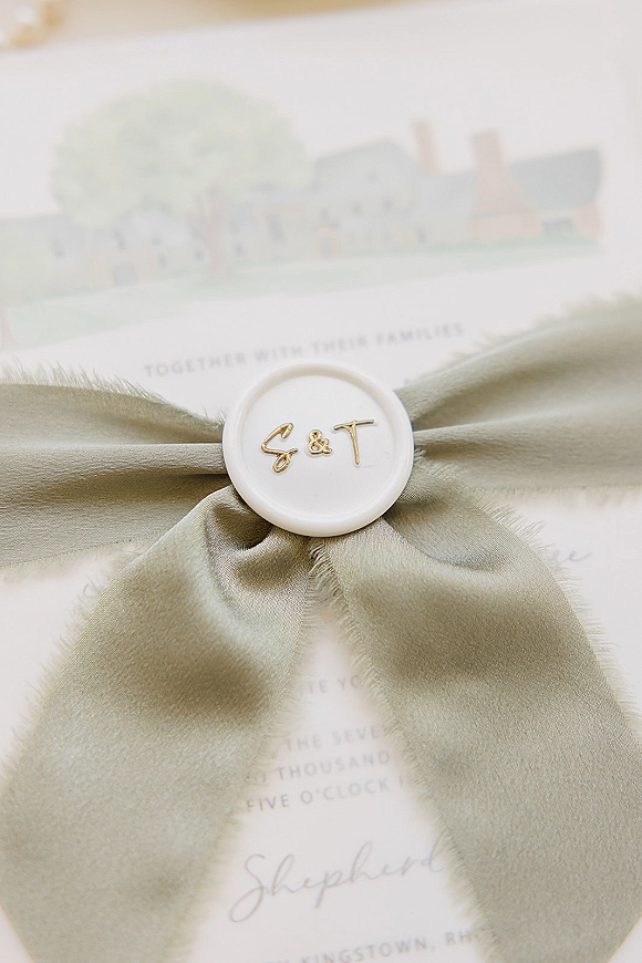 Wedding invitation suite with a sage ribbon bow and custom monogram wax seal, gold initials, and venue card on a neutral surface