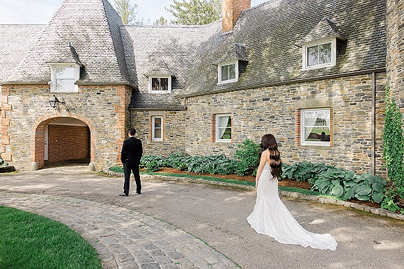 Wedding first look as bride approaches groom from behind, veil and long train flowing on cobblestone drive by stone manor entryway