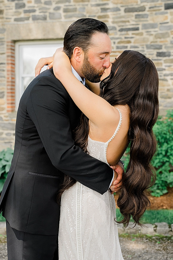 Wedding kiss as bride and groom embrace, her beaded dress straps sparkling, beside a stone wall with window and garden greenery