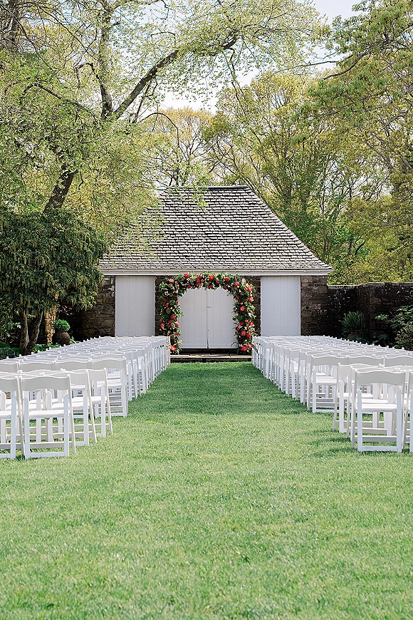Outdoor ceremony setup with white aisle chairs leading to a garden wedding ceremony floral arch on a lawn beside a stone wall and cottage
