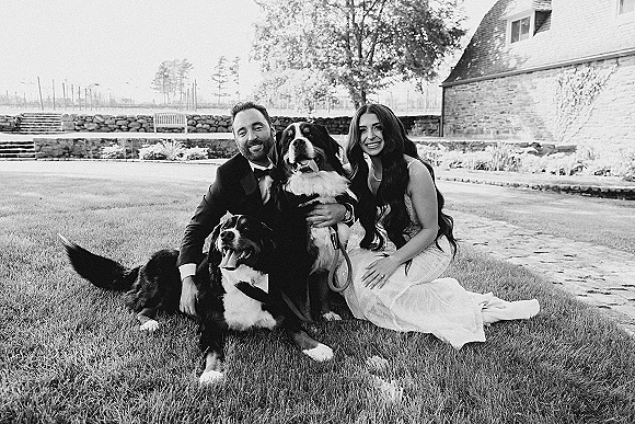Couple portrait of bride in a strapless lace dress and groom in a tuxedo sitting on grass with two leashed dogs by a stone wall