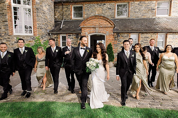 Wedding party portrait with bride and groom with wedding party walking beside a stone building, bride holding a white bouquet on cobblestones