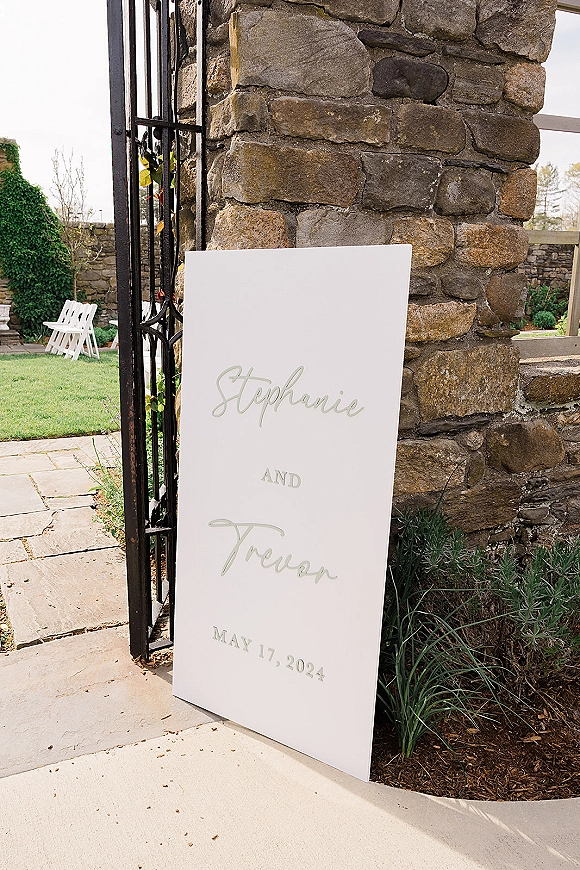 Wedding welcome sign with custom wedding welcome sign calligraphy on a white foam board, leaning by an iron gate near a stone wall walkway