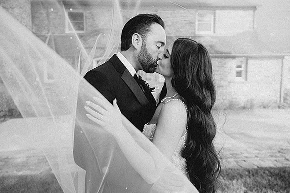 Wedding kiss portrait of bride and groom kissing in a black and white wedding kiss, veil wrapped around them by a stone building