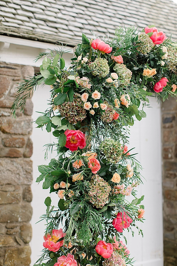 Wedding floral arch with ceremony arch flowers in peonies, hydrangeas, roses and greenery, set against a stone wall with white barn doors