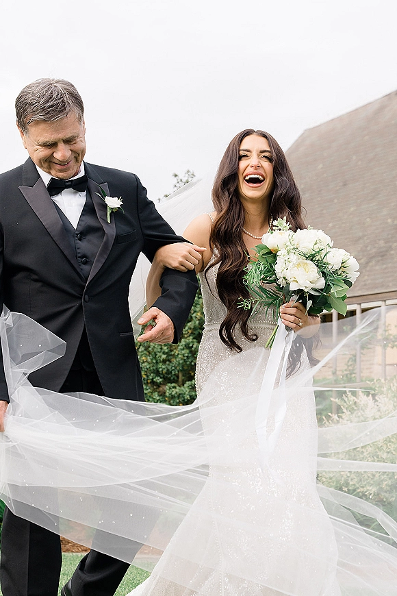 Bride walking down aisle with father, holding a white bridal bouquet as her veil trails, outdoors on a lawn under an overcast sky