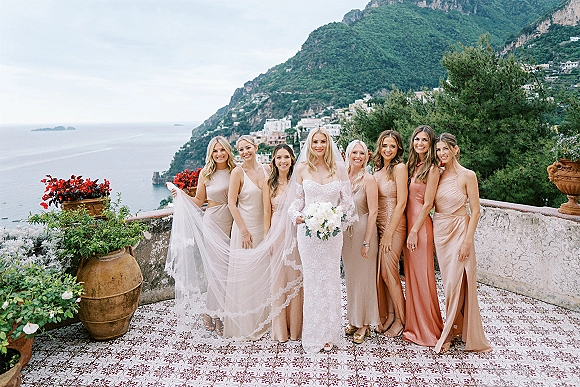 Bridal party portrait of bride with bridesmaids in neutral dresses, bride holding white bouquet on tile terrace with ocean cliffs backdrop.