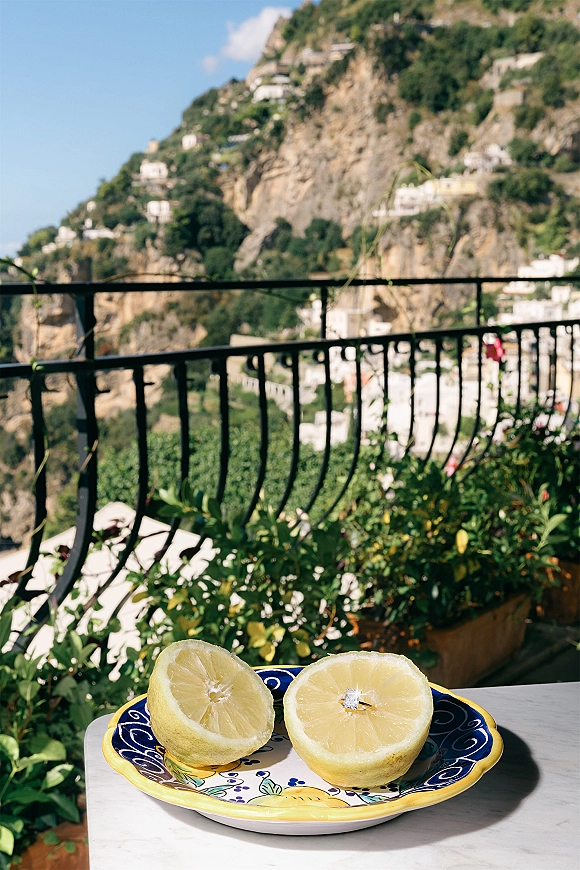 Engagement ring photo of a solitaire diamond nestled in lemon halves on a ceramic plate, set on a sunny cliffside terrace balcony