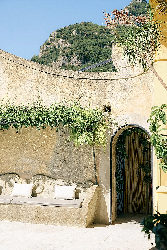 Outdoor lounge seating with built-in bench and neutral cushions, accented by climbing greenery and potted ferns on a sunlit stucco patio