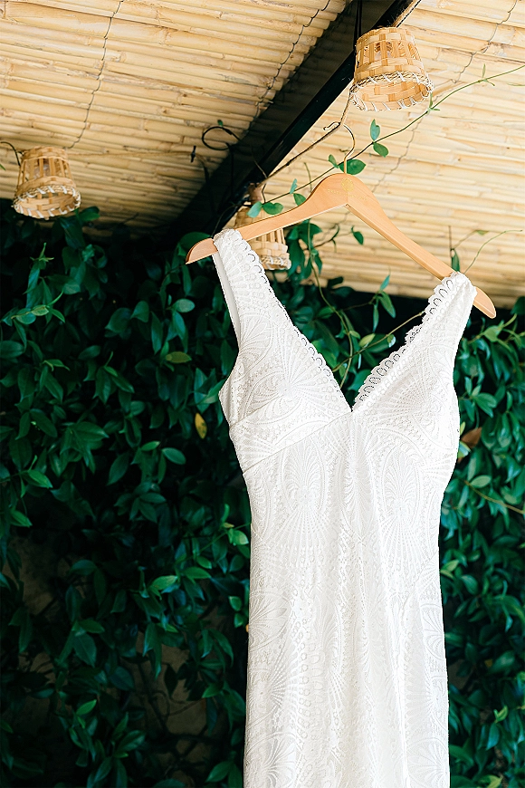 Wedding dress on a wooden hanger, lace wedding dress with V-neck bodice, hanging against a greenery wall under bamboo ceiling lights