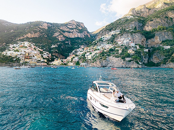 Couple on boat in wedding attire on a motor yacht, bride in dress and groom in suit with sunglasses, ocean cliffs and village behind