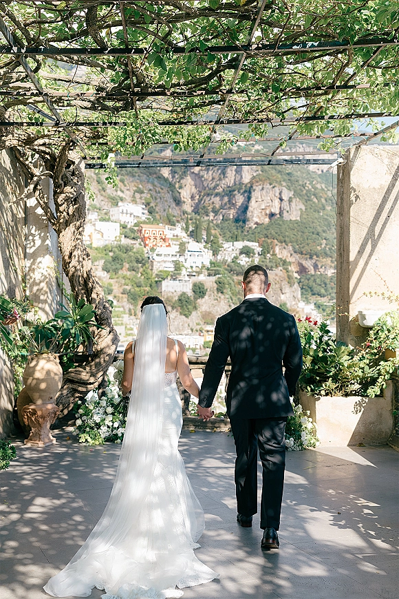 Couple portrait from behind, bride and groom holding hands beneath a vine pergola, her cathedral veil trailing on a stone terrace with cliffs beyond