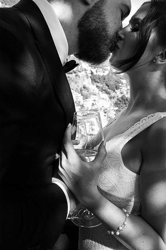 Wedding kiss portrait of bride and groom in black tie, sharing a close up kiss as she holds a champagne flute on a hillside outdoors