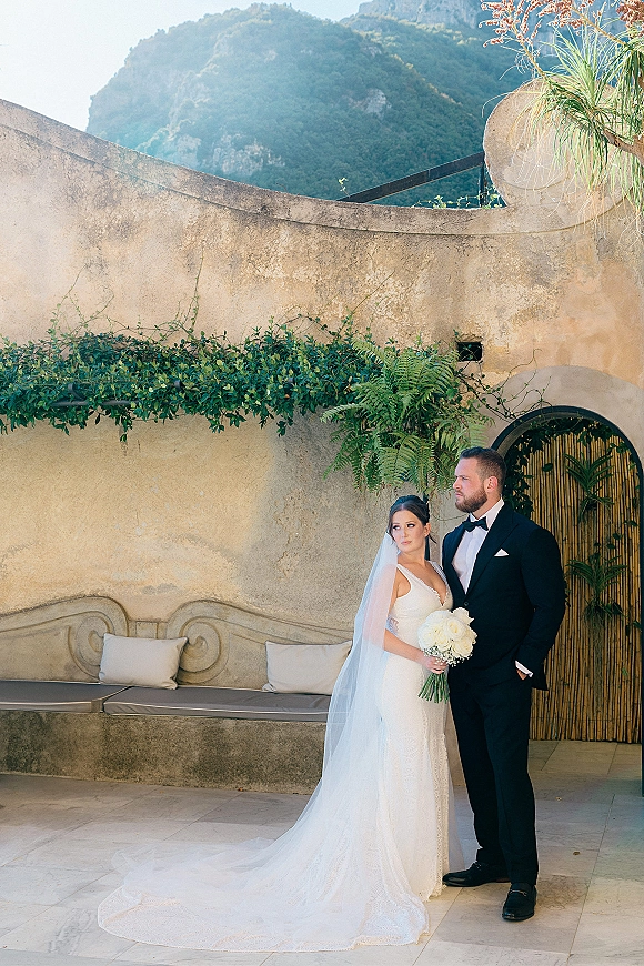 Couple portrait of bride holding a white bouquet beside groom in black tuxedo, long veil flowing by a stucco wall and arched doorway