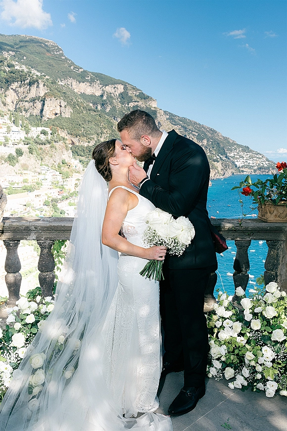Wedding kiss portrait of bride and groom kissing on a terrace, veil blowing in wind, holding white rose bouquet with ocean cliffs behind