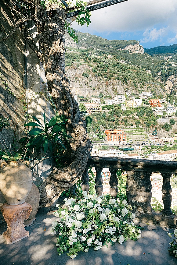 Wedding floral arrangement of white roses and greenery with baby's breath by a stone balustrade on a terrace overlooking a coastal hillside town