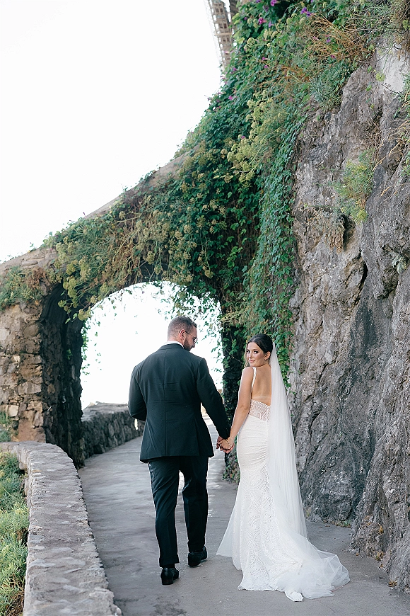 Couple portrait of bride and groom walking away holding hands, her long veil flowing under an ivy-covered stone archway