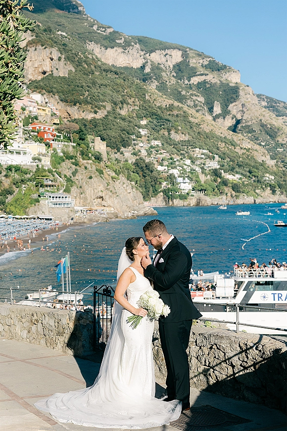 Wedding kiss portrait of bride and groom kissing on a stone terrace with ocean and mountains behind, bride holding a white bouquet and veil