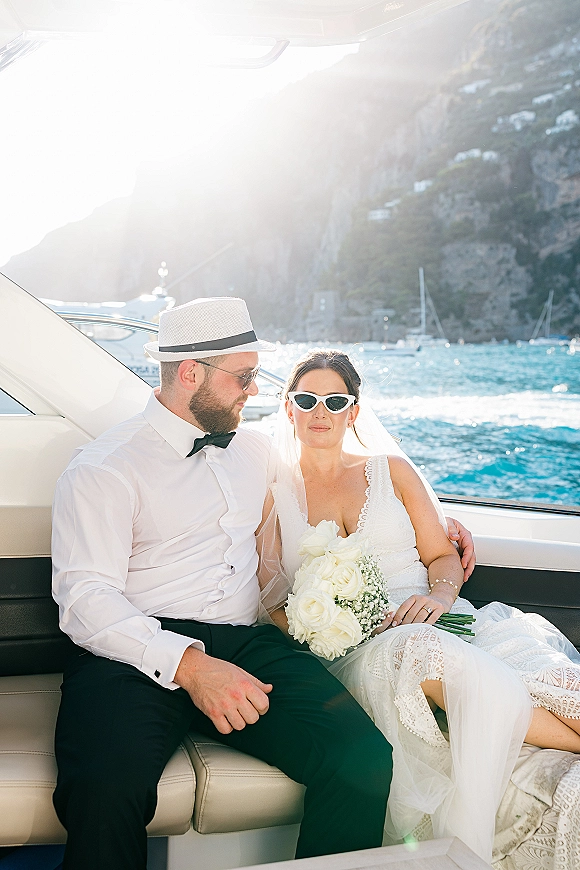 Couple portrait on a wedding on a yacht deck, bride in veil holding white rose bouquet beside groom in fedora, with sailboats and cliffs behind