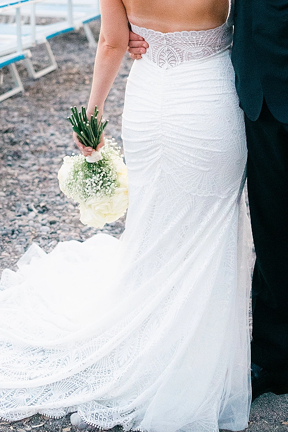 Wedding dress detail showing a strapless lace wedding dress as the bride holds a white rose bouquet with baby’s breath beside a groom in black suit