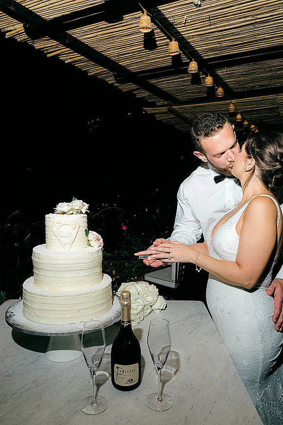 Wedding cake cutting as bride in lace gown and groom in bow tie slice a tiered cake under lantern-lit pergola at night