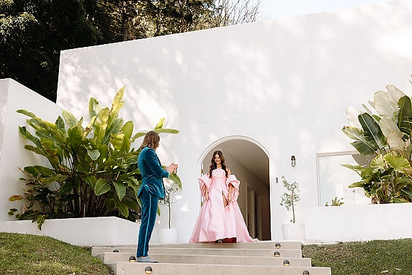 First look moment as groom in teal suit watches bride in a pink gown with statement sleeves and long train on white stucco steps by arched doorway