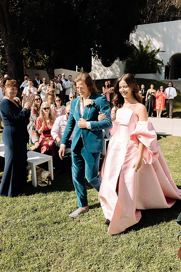 Wedding processional with bride walking down aisle in a strapless pink wedding dress beside partner in teal suit as guests applaud on a sunny lawn