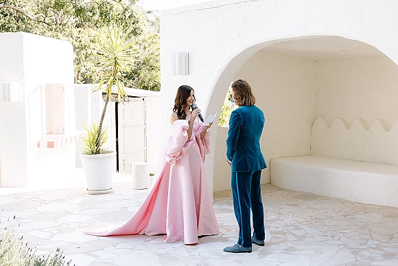 Wedding vows as bride reading vows into a microphone, holding vow paper beside groom in a teal suit under a white stucco arch