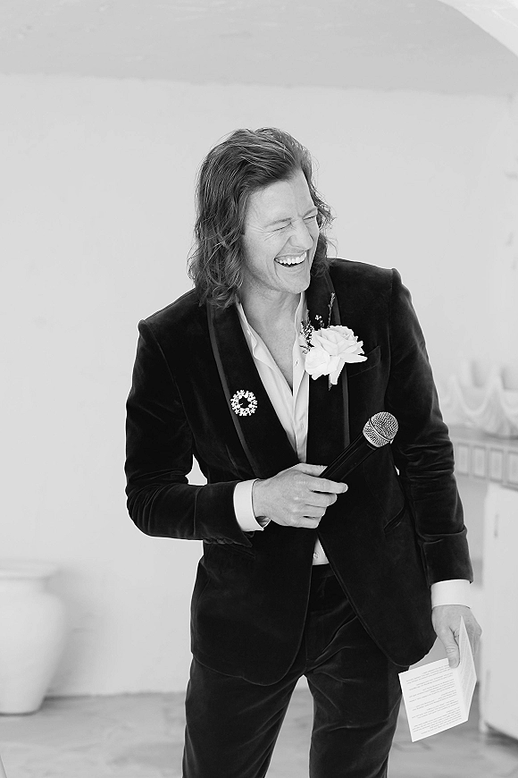 Wedding speech with a handheld microphone as a suited speaker reads paper notes, boutonniere on lapel against a plain indoor wall