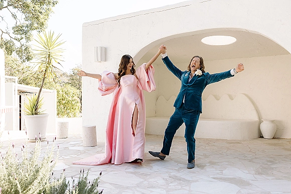 Couple portrait of bride in a pink gown with puff sleeves and slit, and groom in a teal velvet suit, cheering under a white stucco arch courtyard