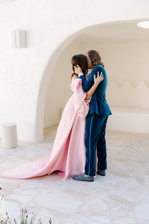 Wedding kiss as the bride in a pink off the shoulder gown with long train and groom in blue suit embrace under a white stucco archway