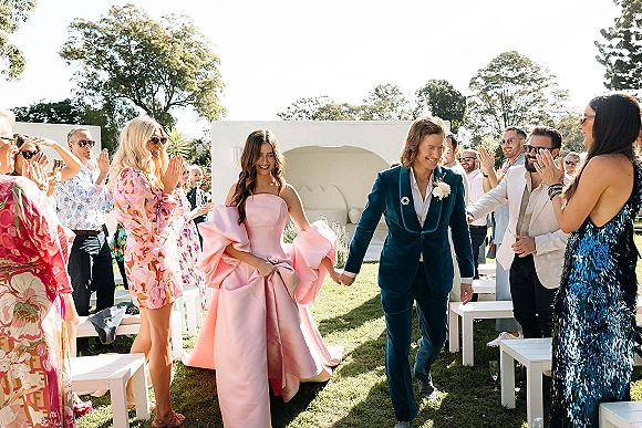 Wedding recessional with bride and groom holding hands, her pink strapless gown train and his teal velvet suit, guests clapping on a lawn aisle