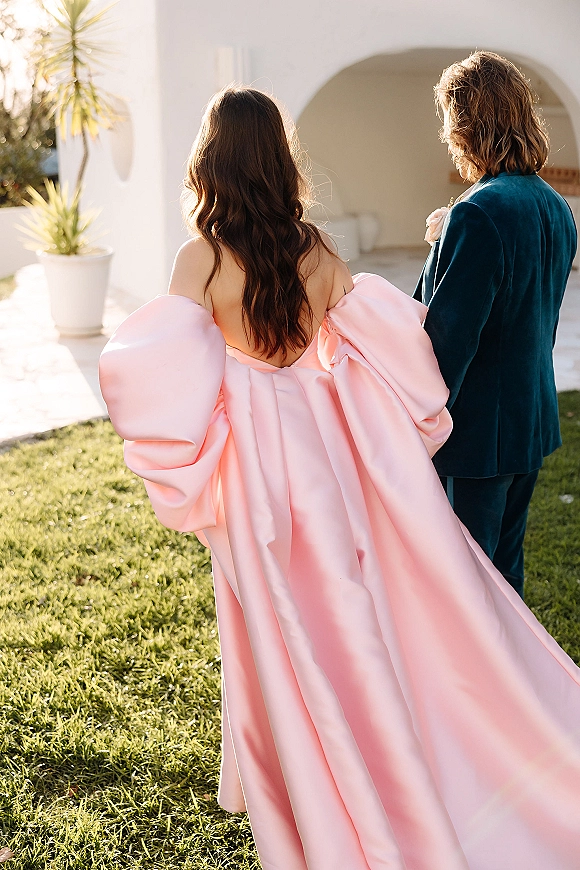 Couple portrait from behind walking away, bride in pink off-the-shoulder satin gown beside groom in velvet suit by a white stucco archway